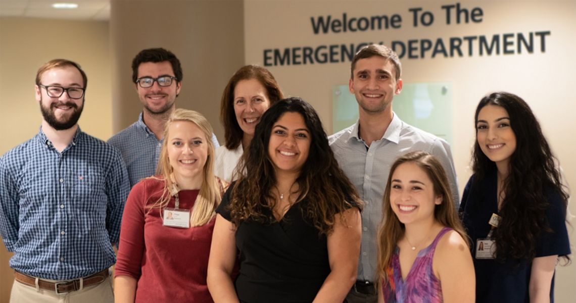 Researchers smiling at the camera standing in front of the Welcome to The Emergency Department sign on a wall